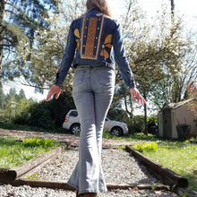 Load image into Gallery viewer, A complete labor of LOVE! This ONE OF A KIND upcycled denim jean jacket features hand appliqued tri-colored earth tone leather flowers throughout the entire jacket along with a &#39;Mayan Temple&#39; inspired design theme on the focal back feature. The unique vintage small crochet trim and center back exquisitely embroidered motif is a delight to the eye having an architectural feel strengthened by the 10mm gold pyramid studs. You will also find our signature hand-embroidered &quot;LOVED&quot; script on the right sleeve.
