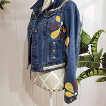 Load image into Gallery viewer, A complete labor of LOVE! This ONE OF A KIND upcycled denim jean jacket features hand appliqued tri-colored earth tone leather flowers throughout the entire jacket along with a &#39;Mayan Temple&#39; inspired design theme on the focal back feature. The unique vintage small crochet trim and center back exquisitely embroidered motif is a delight to the eye having an architectural feel strengthened by the 10mm gold pyramid studs. You will also find our signature hand-embroidered &quot;LOVED&quot; script on the right sleeve.
