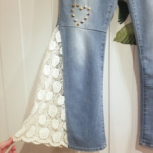 Load image into Gallery viewer, A completely upcycled LEVI denim crop flare jean featuring the sweetest country daisy cotton crochet lace with scalloping along the edges set in a unique asymmetrical detailing, gold + silver hand appliqued studded heart motifs, a one sided cotton eyelet trimmed side seam and 2 of our hand-embroidered LOVED scripts in soft pastel tones of gold, ivory and pink. Scoop up these beauties and feel loved everyday!
