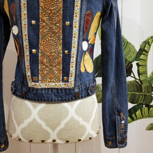 Load image into Gallery viewer, A complete labor of LOVE! This ONE OF A KIND upcycled denim jean jacket features hand appliqued tri-colored earth tone leather flowers throughout the entire jacket along with a &#39;Mayan Temple&#39; inspired design theme on the focal back feature. The unique vintage small crochet trim and center back exquisitely embroidered motif is a delight to the eye having an architectural feel strengthened by the 10mm gold pyramid studs. You will also find our signature hand-embroidered &quot;LOVED&quot; script on the right sleeve.
