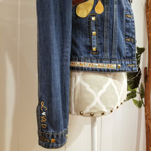 Load image into Gallery viewer, A complete labor of LOVE! This ONE OF A KIND upcycled denim jean jacket features hand appliqued tri-colored earth tone leather flowers throughout the entire jacket along with a &#39;Mayan Temple&#39; inspired design theme on the focal back feature. The unique vintage small crochet trim and center back exquisitely embroidered motif is a delight to the eye having an architectural feel strengthened by the 10mm gold pyramid studs. You will also find our signature hand-embroidered &quot;LOVED&quot; script on the right sleeve.
